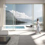 A modern living room with floor-to-ceiling aluminium sliding doors opening to a stunning mountain and lake view. A woman in a white dress walks gracefully through the open doors, emphasizing the seamless indoor-outdoor connection and elegance of the space.